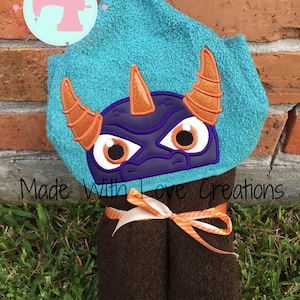 May include: A turquoise hooded towel with an embroidered purple dragon face with orange horns and white eyes. The towel is tied with a white ribbon with orange stripes. "Made With Love Creations" is written on the towel.
