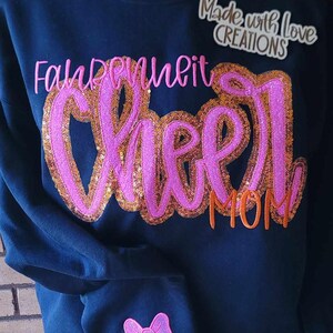 May include: A black sweatshirt with the words "Fall in love with cheer mom" in pink and orange glitter lettering. The sweatshirt also has a pink glitter bow with the name "Camryn" in orange lettering on the sleeve.