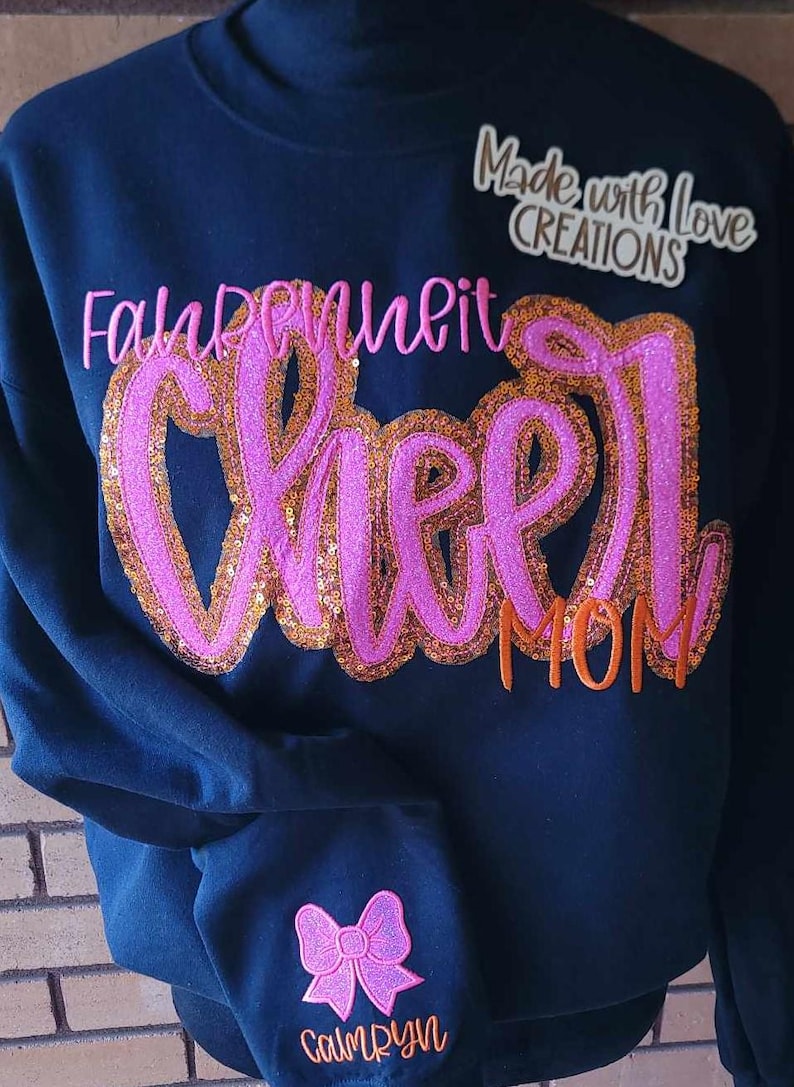 May include: A black sweatshirt with the words "Fall in love with cheer mom" in pink and orange glitter lettering. The sweatshirt also has a pink glitter bow with the name "Camryn" in orange lettering on the sleeve.