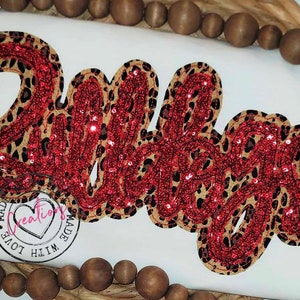 May include: A red and brown sequined appliqu&eacute; of the word "Babe" on a white background. The appliqu&eacute; is made with a leopard print fabric and red sequins.