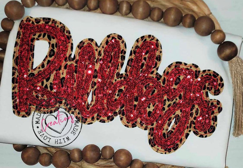 May include: A red and brown sequined appliqu&eacute; of the word "Babe" on a white background. The appliqu&eacute; is made with a leopard print fabric and red sequins.