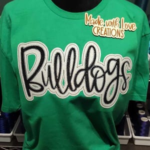 May include: Green t-shirt with a white and black "Bulldogs" design. The shirt has a small brown tag that says "Made with Love Creations".