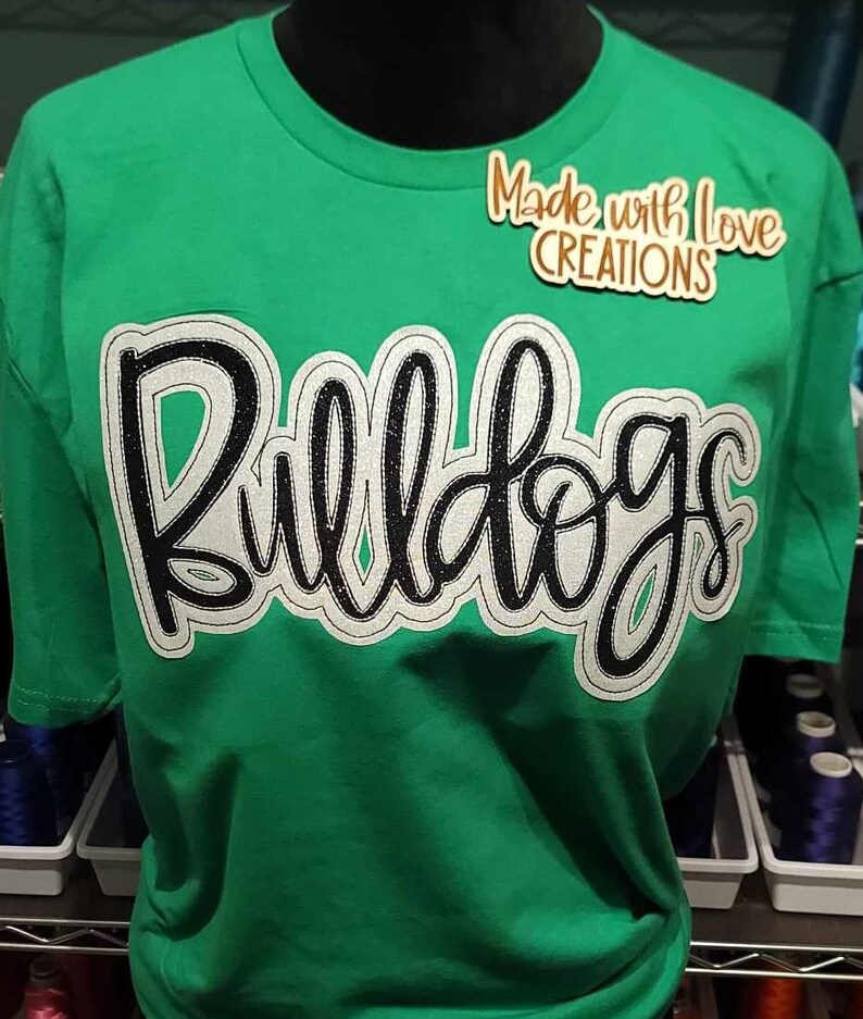 May include: Green t-shirt with a white and black "Bulldogs" design. The shirt has a small brown tag that says "Made with Love Creations".