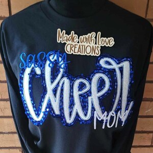 May include: Black sweatshirt with blue and white sequined lettering that reads "Sassy Cheer Mom" and a brown label that reads "Made with Love Creations".
