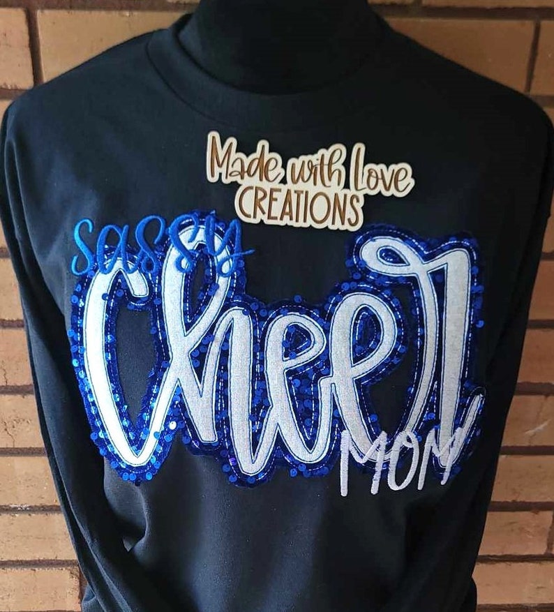 May include: Black sweatshirt with blue and white sequined lettering that reads "Sassy Cheer Mom" and a brown label that reads "Made with Love Creations".