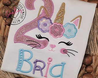 May include: White t-shirt with a pink glittery number 2, cat face, and the name "Bria" in blue and pink embroidery. The cat face has black whiskers and closed eyes, with floral and gold accents. The shirt is on a woven background.