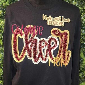 May include: Black long-sleeve sweatshirt with gold and burgundy glitter lettering that reads "Colonie Cheer Mom" and a small gold label that reads "Made with Love Creations".