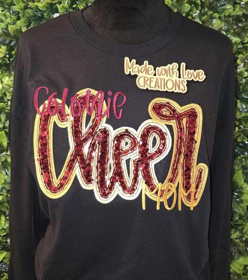 May include: Black long-sleeve sweatshirt with gold and burgundy glitter lettering that reads "Colonie Cheer Mom" and a small gold label that reads "Made with Love Creations".