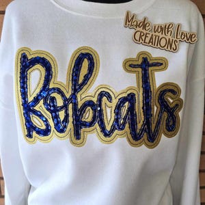 May include: White sweatshirt with the word "Bobcats" in blue sequins outlined in gold. The sweatshirt has a crew neck and a small wooden tag that says "Made with Love CREATIONS".