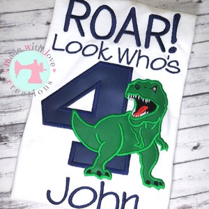 May include: A white t-shirt with the text "Roar! Look who's 4 John" in blue embroidery. A green dinosaur with a blue outline is embroidered next to the number 4.