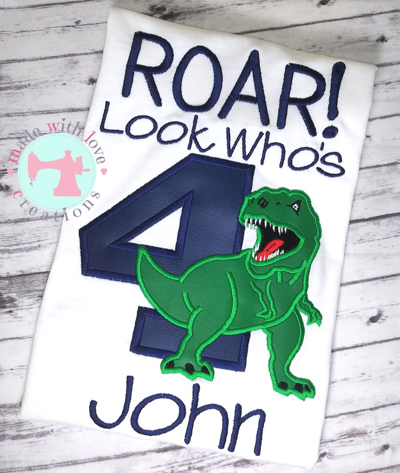 May include: A white t-shirt with the text "Roar! Look who's 4 John" in blue embroidery. A green dinosaur with a blue outline is embroidered next to the number 4.