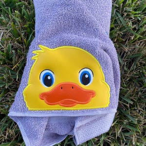 May include: A purple hooded towel with a yellow embroidered duck face. The duck has blue eyes and an orange beak.