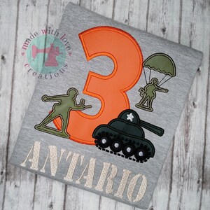 May include: A gray t-shirt with an orange number 3, a green tank, a green soldier with a parachute, and a green soldier holding a gun. The text "ANTARIO" is embroidered in beige below the design.