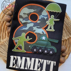 May include: A black t-shirt with a camouflage number eight, a green tank, a soldier with a parachute, and another soldier holding a gun. The text "EMMETT" is embroidered in white below the design.