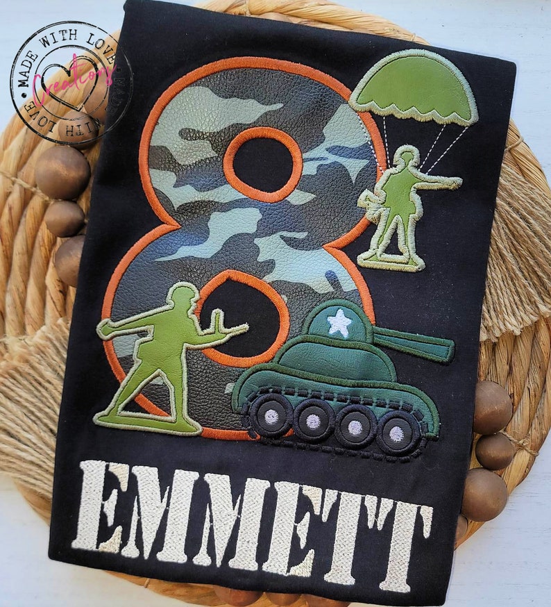 May include: A black t-shirt with a camouflage number eight, a green tank, a soldier with a parachute, and another soldier holding a gun. The text "EMMETT" is embroidered in white below the design.