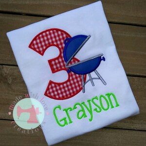 May include: A white t-shirt with a red and white gingham number 3, a blue grill, and the name Grayson in green text.