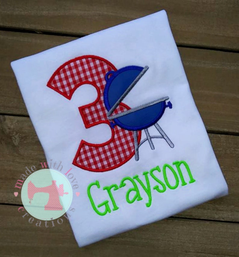 May include: A white t-shirt with a red and white gingham number 3, a blue grill, and the name Grayson in green text.