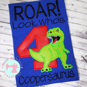 May include: Blue t-shirt with the text "Roar! Look who's 4 Coopersaurus" in black embroidery. A green dinosaur with a red number 4 is embroidered on the shirt.