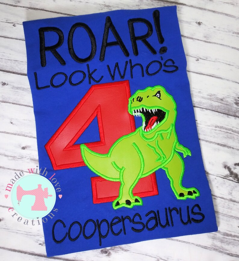 May include: Blue t-shirt with the text "Roar! Look who's 4 Coopersaurus" in black embroidery. A green dinosaur with a red number 4 is embroidered on the shirt.