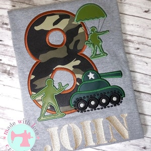 May include: A gray t-shirt with a camouflage number eight, a green tank, a green soldier with a parachute, and a green soldier. The text "JOHN" is embroidered in gold below the design.