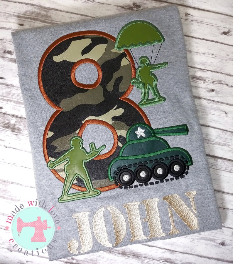 May include: A gray t-shirt with a camouflage number eight, a green tank, a green soldier with a parachute, and a green soldier. The text "JOHN" is embroidered in gold below the design.
