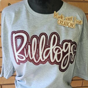 May include: Grey t-shirt with a burgundy glitter design that says "Bulldogs". The shirt also has a small wooden tag that says "Made with Love Creations".
