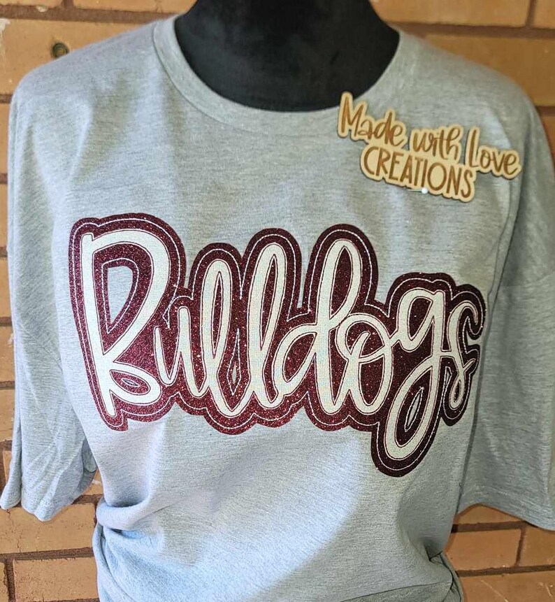 May include: Grey t-shirt with a burgundy glitter design that says "Bulldogs". The shirt also has a small wooden tag that says "Made with Love Creations".