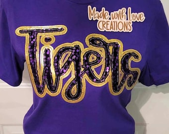 May include: Purple short-sleeved t-shirt with a gold and purple sequined "Tigers" design. The shirt has the text "Made with Love Creations" on the top right corner.