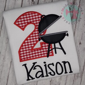 May include: Red and white gingham number two with a black grill and the name Kaison in black embroidery on a white t-shirt.