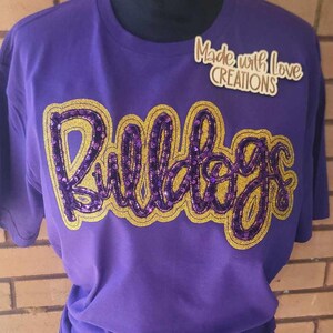 May include: Purple t-shirt with a gold and purple sequined "Bulldogs" design. The shirt has a crew neck and short sleeves. The text "Made with Love Creations" is visible in the top right corner of the image.