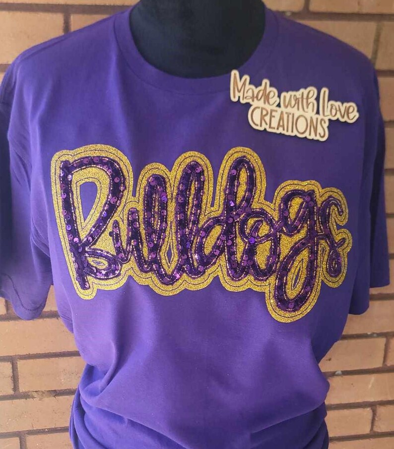 May include: Purple t-shirt with a gold and purple sequined "Bulldogs" design. The shirt has a crew neck and short sleeves. The text "Made with Love Creations" is visible in the top right corner of the image.