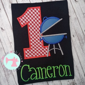 May include: A black t-shirt with a red and white gingham number one, a blue grill, and the name "Cameron" in green embroidery.