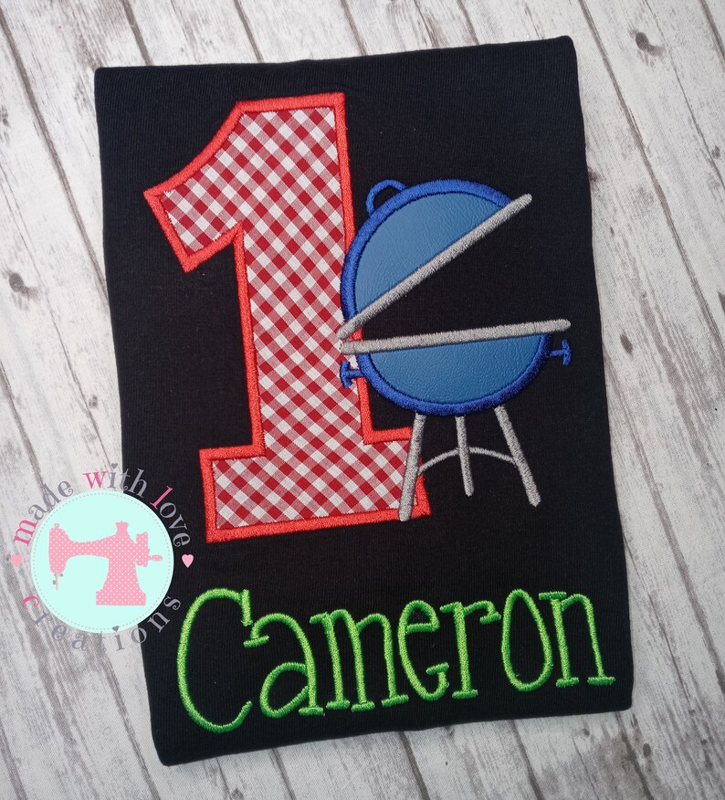 May include: A black t-shirt with a red and white gingham number one, a blue grill, and the name "Cameron" in green embroidery.