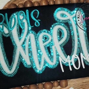 May include: A black t-shirt with the words "Girls Cheer Mom" in white and turquoise sequins.