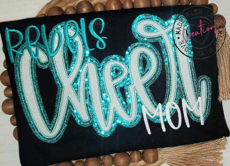 May include: A black t-shirt with the words "Girls Cheer Mom" in white and turquoise sequins.