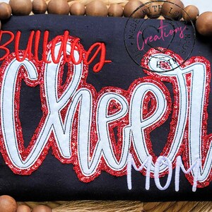 May include: A black t-shirt with red and white sequined lettering that reads "Bulldog Cheer Mom".