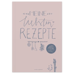 Könnte beinhalten: Ein rosa Einband für ein Rezeptbuch mit dem Titel "Meine liebsten Rezepte" in blauer Schrift. Der Einband zeigt außerdem eine blaue Illustration von Küchenutensilien und Kräutern.