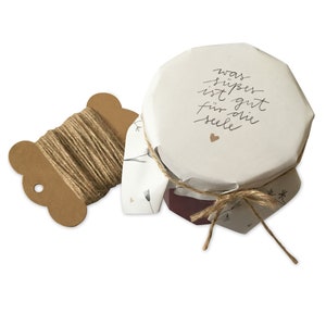 Op de afbeelding: Een glazen pot met een wit deksel dat is ingepakt in bruin papier en vastgebonden met touw. Het deksel heeft de tekst "was süpes ist gut für die Seele" erop.