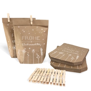 Op de afbeelding: Kraftpapieren cadeauzakjes met witte sneeuwvlok- en bloemmotieven, en de tekst "FROHE Weihnachten". De zakjes zijn afgesloten met houten wasknijpers. Een stapel bijpassende papieren labels en een stapel wasknijpers zijn ook zichtbaar.
