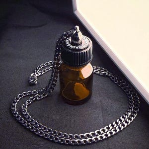 May include: A small, brown glass bottle with a black cap and a silver chain necklace. The bottle is surrounded by the chain, which is made of small, interlocking links. The cap has the text "CHAINS FOR LIFE" on it.
