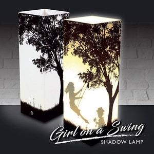 May include: Two rectangular shadow lamps with a silhouette design. One lamp is unlit, the other glows with a warm light, showing a girl on a swing under a tree. The lamps have a black base and a white top. The text "Girl on a Swing SHADOW LAMP" is at the bottom.