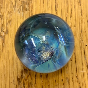 May include: A round glass paperweight with a swirling blue, green, and purple design. The glass contains gold glitter and small air bubbles.