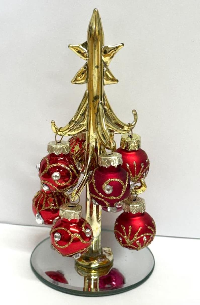 May include: A small Christmas tree ornament. The tree is made of gold-coloured metal, topped with a star, and holds several small, red, round baubles with gold and rhinestone detailing. It sits on a mirrored base.