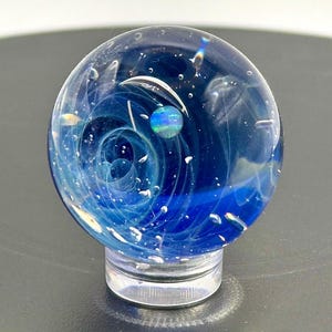 Puede incluir: Una canica de vidrio transparente con un diseño en espiral azul y blanco, que recuerda a una galaxia. Tiene un pequeño orbe iridiscente y descansa sobre un soporte transparente. El diseño crea una representación artística en miniatura del espacio.