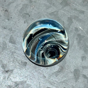 May include: A round glass paperweight with swirling black, white, and blue patterns. The center of the paperweight is a dark black circle.