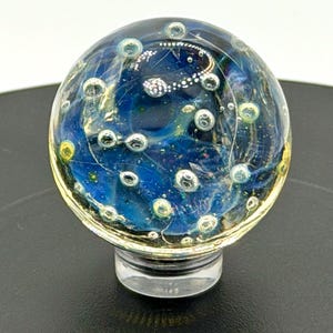 Può includere: Un fermacarte sferico in vetro trasparente con interno blu e oro, con bolle incorporate e un design a spirale. Il fermacarte poggia su una base cilindrica trasparente. Il numero "102" è visibile in basso.