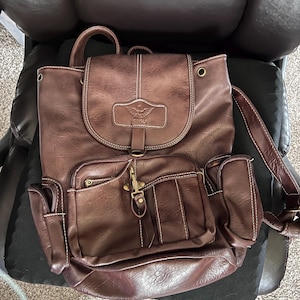 Puede incluir: Mochila de cuero marrón con múltiples bolsillos y cierre de solapa. Cuenta con costuras blancas y un cierre dorado. La mochila tiene un asa superior y correas ajustables. La palabra "YONGLE" es visible en la solapa delantera.