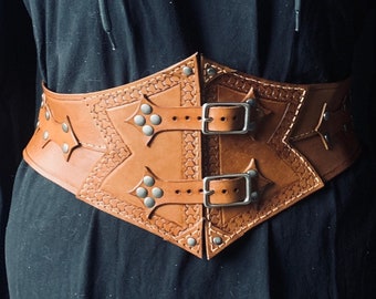 May include: A brown leather belt with silver buckles and studs. The belt has a decorative design with pointed shapes and is worn over a black hooded garment.