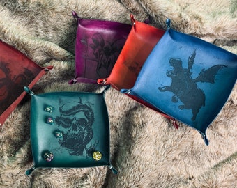 May include: Five leather dice trays with different designs. The trays are in various colors, including red, blue, green, and purple. Each tray has a different design, including a skull, a dragon, and a hippogriff. The trays are on a faux fur surface.