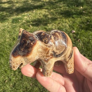 May include: A small, carved animal figurine made of brown and white banded onyx. The figurine is shaped like a pig or a bear and has a smooth, polished finish.